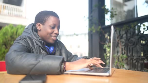 Glücklich lächelnde schwarzhaarige Studentin in schwarzer Daunenjacke mit blauen Kopfhörern, die im Straßencafé am Laptop lernt. Vielfalt. Fernarbeit, Fernstudium. — Stockvideo