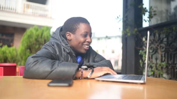 Glücklich lächelnde afrikanische Studentin mit schwarzem Kurzhaarschnitt in schwarzer Daunenjacke und blauen Kopfhörern unterhält sich auf dem Laptop im Straßencafé. Vielfalt. Fernarbeit, Fernstudium. — Stockvideo