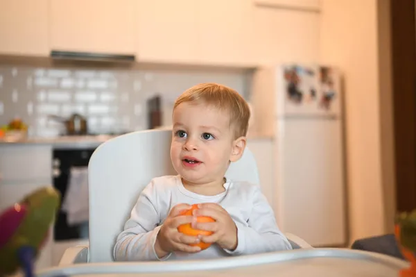 Poco feliz bebé lindo niño rubio sentado en silla de bebé jugando con naranja. Bebé expresiones faciales en el interior de la casa en la cocina interior con comida. Comer sano concepto de infancia familiar feliz. —  Fotos de Stock