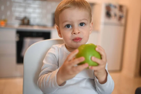 Poco feliz bebé lindo niño rubio sentado en silla de bebé jugando con manzana. Bebé expresiones faciales en el interior de la casa en la cocina interior con comida. Comer sano concepto de infancia familiar feliz. —  Fotos de Stock