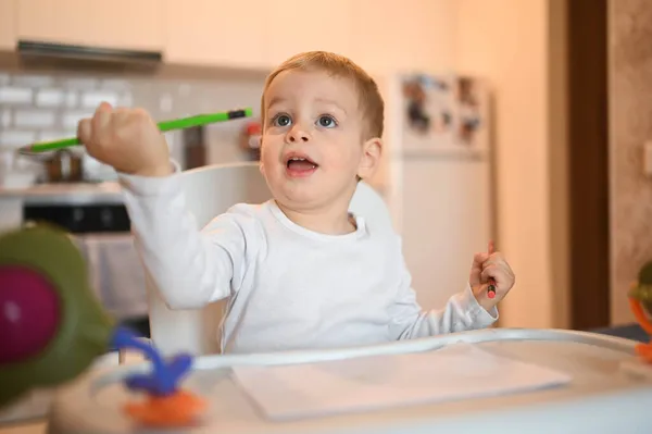Pequeño bebé lindo niño rubio sentado en silla de bebé aprender dibujar con lápices de color. Bebé expresiones faciales en el interior de la casa en la cocina interior con juguetes. Saludable concepto de infancia familiar feliz. —  Fotos de Stock