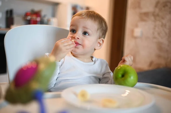 Poco feliz bebé lindo niño rubio sentado en silla de bebé jugando con manzana. Bebé expresiones faciales en el interior de la casa en la cocina interior con comida. Comer sano concepto de infancia familiar feliz. —  Fotos de Stock