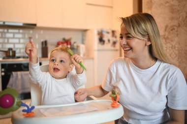 Küçük tatlı bebek bebek sarışın bebek bebek sandalyede oturup resim yapmayı öğreniyor. Güzel anne ve oğul evde oyuncaklarla birlikte vakit geçiriyorlar. Sağlıklı mutlu aile çocukluğu kavramı.