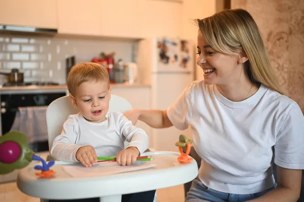 Kleine schattige baby peuter jongen blond zittend op kinderstoel leren tekenen. Mooie jonge moeder en zoon spelen tijd samen binnen met speelgoed. Gezonde gelukkige familie kindertijd concept. — Stockfoto