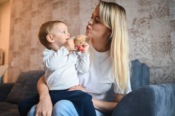 Mały śliczny chłopczyk blondyn siedzący na ramionach matek. Piękna młoda mama i syn bawią się w domu z zabawkami. Zdrowa koncepcja szczęśliwego dzieciństwa. — Zdjęcie stockowe