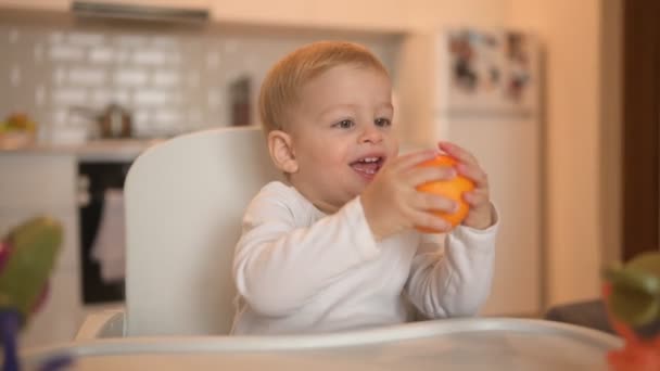 Beetje vrolijk schattig baby peuter jongen blond zittend op kinderstoel spelen met oranje. Baby gezichtsuitdrukkingen binnen thuis keuken interieur met voedsel. Gezond eten gelukkig gezin kindertijd concept. — Stockvideo