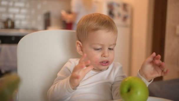 Pouco feliz bebê bonito criança menino loiro sentado na cadeira do bebê brincando com a maçã. Expressões faciais do bebê dentro de casa cozinha interior com comida. Alimentação saudável conceito de infância familiar feliz. — Vídeo de Stock