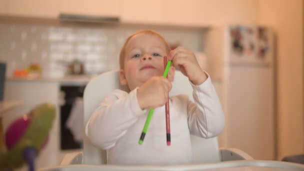 Pouco bonito bebê criança menino loiro sentado na cadeira de bebê aprendendo desenhar com lápis de cor. Expressões faciais do bebê dentro de casa cozinha interior com brinquedos. Conceito de infância familiar feliz saudável. — Vídeo de Stock
