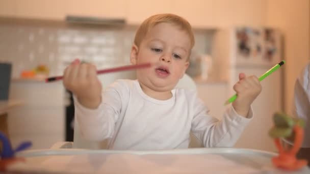 Liten söt bebis barn pojke blond sitter på barnstol lära rita med färgpennor. Baby ansiktsuttryck inomhus kök interiör med leksaker. Friska lycklig familj barndom koncept. — Stockvideo