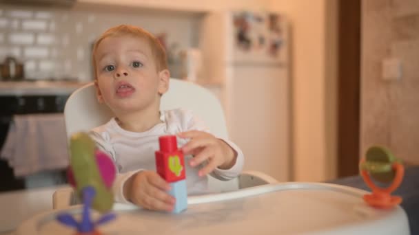 Malá šťastná roztomilá batole chlapec blondýnka sedí na dětské židli hrát s konstruktérem. Dětské výrazy obličeje v interiéru domácí kuchyně s hračkami. Zdravé šťastné rodinné dětství koncept. — Stock video