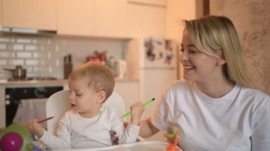 Küçük tatlı bebek bebek sarışın bebek bebek sandalyede oturup resim yapmayı öğreniyor. Güzel anne ve oğul evde oyuncaklarla birlikte vakit geçiriyorlar. Sağlıklı mutlu aile çocukluğu kavramı.
