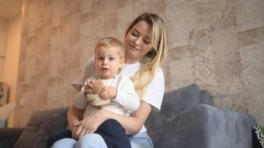 Küçük tatlı bebek sarışının annesinin kollarında oturması. Güzel anne ve oğul evde oyuncaklarla birlikte vakit geçiriyorlar. Sağlıklı mutlu aile çocukluğu kavramı.