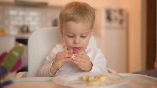 Mały szczęśliwy słodkie dziecko maluch chłopiec blondynka siedzi na foteliku bawiąc się z bananem. Wyrazy twarzy dziecka wewnątrz w domu kuchnia wnętrze z jedzeniem. Zdrowe jedzenie koncepcja szczęśliwego dzieciństwa rodziny. — Wideo stockowe