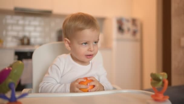 Liten glad söt bebis barn pojke blond sitter på barnstol leker med orange. Baby ansiktsuttryck inomhus kök interiör med mat. Friska äta lycklig familj barndom koncept. — Stockvideo