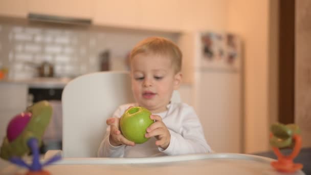 Poco feliz bebé lindo niño rubio sentado en silla de bebé jugando con manzana. Bebé expresiones faciales en el interior de la casa en la cocina interior con comida. Comer sano concepto de infancia familiar feliz. — Vídeos de Stock