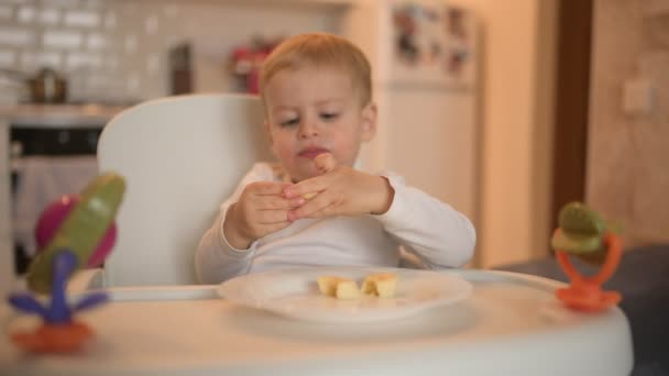 Liten glad söt liten pojke blond sitter på barnstol leker med banan. Baby ansiktsuttryck inomhus kök interiör med mat. Friska äta lycklig familj barndom koncept. — Stockvideo