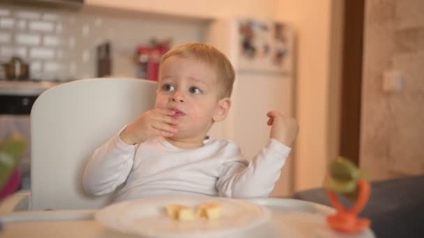 Malá šťastná roztomilá batole kluk blondýnka sedí na dětské židli hrát s banánem. Dětské výrazy obličeje v interiéru domácí kuchyně s jídlem. Zdravé stravování šťastný rodinný dětství koncept. — Stock video