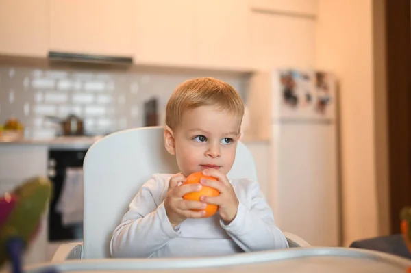 Poco feliz bebé lindo niño rubio sentado en silla de bebé jugando con naranja. Bebé expresiones faciales en el interior de la casa en la cocina interior con comida. Comer sano concepto de infancia familiar feliz. —  Fotos de Stock
