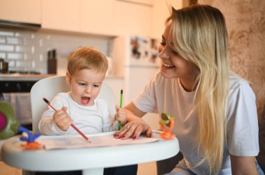 Küçük tatlı bebek bebek sarışın bebek bebek sandalyede oturup resim yapmayı öğreniyor. Güzel anne ve oğul evde oyuncaklarla birlikte vakit geçiriyorlar. Sağlıklı mutlu aile çocukluğu kavramı.