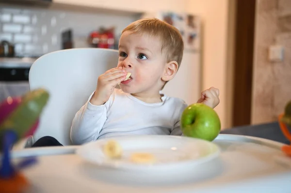Poco feliz bebé lindo niño rubio sentado en silla de bebé jugando con manzana. Bebé expresiones faciales en el interior de la casa en la cocina interior con comida. Comer sano concepto de infancia familiar feliz. —  Fotos de Stock