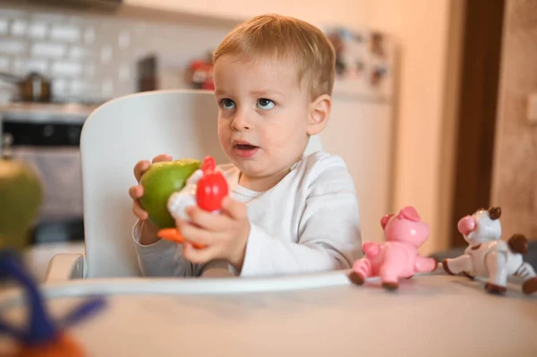 Poco feliz bebé lindo niño rubio sentado en silla de bebé jugando con manzana. Bebé expresiones faciales en el interior de la casa en la cocina interior con comida. Comer sano concepto de infancia familiar feliz. —  Fotos de Stock