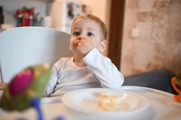 Poco feliz bebé lindo niño rubio sentado en silla de bebé jugando con plátano. Bebé expresiones faciales en el interior de la casa en la cocina interior con comida. Comer sano concepto de infancia familiar feliz. —  Fotos de Stock