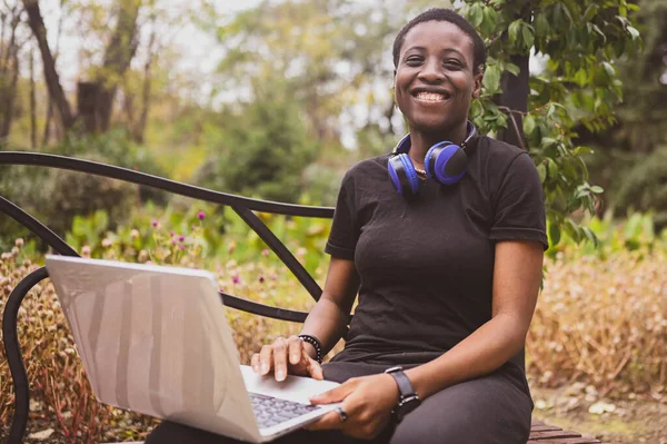 Lachend vrolijk Afrikaans zwart kortharige vrouw studente afro haar met blauwe koptelefoon die online studeert op laptop computer in de zomer groen park. Verscheidenheid. Afstandswerk, afstandsonderwijs. — Stockfoto