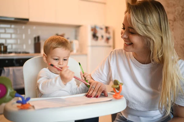 Kleine schattige baby peuter jongen blond zittend op kinderstoel leren tekenen. Mooie jonge moeder en zoon spelen tijd samen binnen met speelgoed. Gezonde gelukkige familie kindertijd concept. — Stockfoto