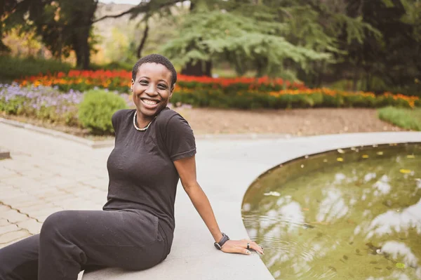 Aantrekkelijke vrolijke glimlach jonge natuurlijke schoonheid kortharige Afrikaanse vrouw dragen totaal zwart lachen zitten aan de rand van een fontein in de natuur zomer park. — Stockfoto