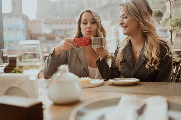 Retrato de cerca de dos amigas con trajes estrictos riendo bebiendo café y vino en la terraza al aire libre en la cafetería de la calle de verano en los edificios de fondo de la antigua ciudad de Tiflis, Georgia —  Fotos de Stock