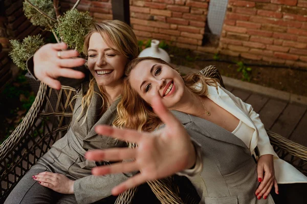 Potret close-up dari dua teman perempuan dengan setelan abu-abu yang ketat tertawa dan menutup wajah dengan tangan dari kamera sambil duduk di kursi wicker di teras luar — Stok Foto