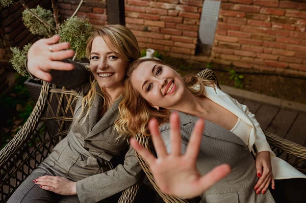 Potret close-up dari dua teman perempuan dengan setelan abu-abu yang ketat tertawa dan menutup wajah dengan tangan dari kamera sambil duduk di kursi wicker di teras luar — Stok Foto