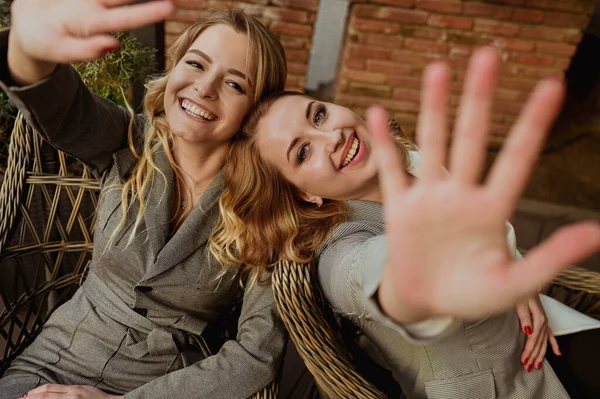 Potret close-up dari dua teman perempuan dengan setelan abu-abu yang ketat tertawa dan menutup wajah dengan tangan dari kamera sambil duduk di kursi wicker di teras luar — Stok Foto