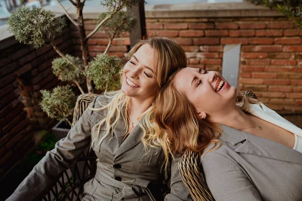 Potret close-up dari dua teman perempuan dengan setelan abu-abu yang ketat tertawa sambil duduk di kursi wicker di teras luar di kafe jalan musim panas. — Stok Foto