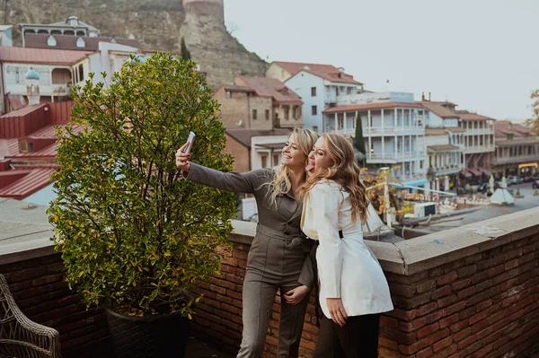 Katı takım elbiseli iki kadının yakın plan portresi, Georgia 'daki Old Tiflis şehrinin arka planındaki yazlık kafede selfie çekerken gülüyor..