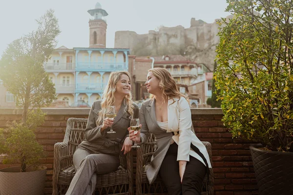 Foto close-up dari dua teman perempuan dengan pakaian ketat tertawa sambil minum anggur di teras luar di kafe musim panas di bangunan latar belakang kota Tbilisi Lama, Georgia. — Stok Foto