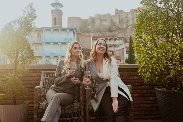 Katı takım elbiseli iki kadının yakın plan portresi. Georgia 'daki Old Tiflis şehrinin arka planındaki yazlık kafede şarap içerek gülüyorlar..