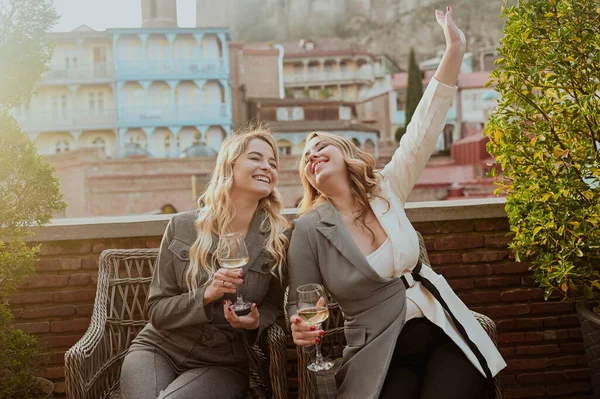 Foto close-up dari dua teman perempuan dengan pakaian ketat tertawa sambil minum anggur di teras luar di kafe musim panas di bangunan latar belakang kota Tbilisi Lama, Georgia. — Stok Foto