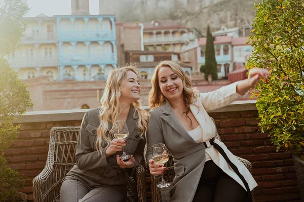 Zbliżenie portret dwóch przyjaciółek w surowych garniturach śmiejących się z wina na tarasie przed kawiarnią przy letniej ulicy na tle budynków Starego Tbilisi, Gruzja. — Zdjęcie stockowe