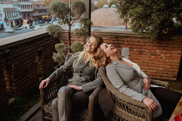 Potret close-up dari dua teman perempuan dengan setelan abu-abu yang ketat tertawa sambil duduk di kursi wicker di teras luar di kafe jalan musim panas. — Stok Foto