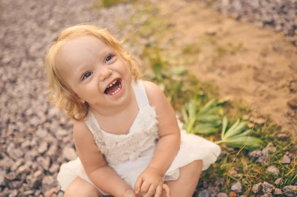 夏天的时候，一个穿着白衣、脸上沾满泥巴的可爱的金发小女孩，坐在外面街上的地上。宝宝笑了。快乐健康童年的概念. — 图库照片