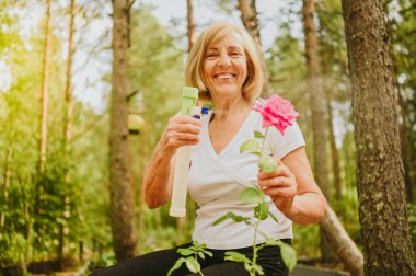 Yaşlı, kıdemli bahçıvan kadın, kır bahçesinde çiçeklere bakan kırsal alanda, su tozu kullanarak çiçek açan bitkileri ilaçlıyor. Çiftçilik, bahçıvanlık, tarım, emekli yaşlılar..