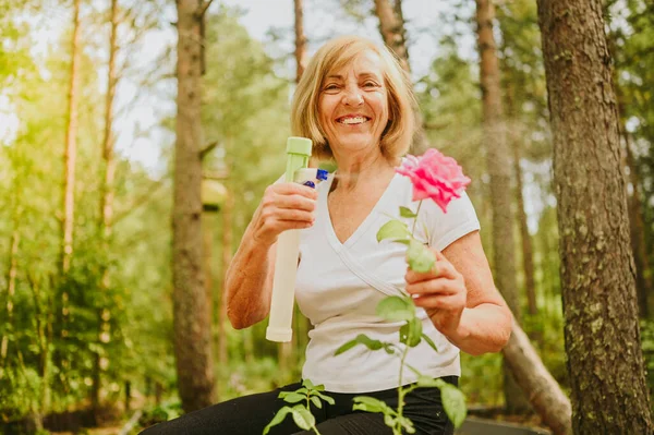 Yaşlı, kıdemli bahçıvan kadın, kır bahçesinde çiçeklere bakan kırsal alanda, su tozu kullanarak çiçek açan bitkileri ilaçlıyor. Çiftçilik, bahçıvanlık, tarım, emekli yaşlılar..