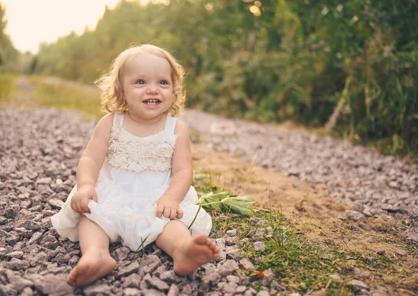 Beyaz elbiseli ve yüzünde çamurla küçük sevimli sarışın kız çocuğu yazın dışarıdaki caddede oturuyor. Bebek gülüyor. Mutlu sağlıklı çocukluk kavramı.