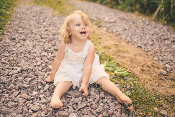 夏天的时候，一个穿着白衣、脸上沾满泥巴的可爱的金发小女孩，坐在外面街上的地上。宝宝笑了。快乐健康童年的概念. — 图库照片