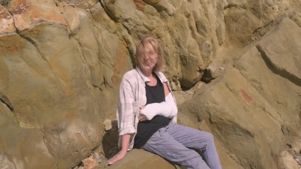 Seniors souriant femme âgée se détendre assis sur la côte rocheuse dans la journée venteuse souffrant de douleurs articulaires. Blessure à la main. Vieille dame poignet en gypse bandage fronde se sentir blessé à l'extérieur en été. — Video
