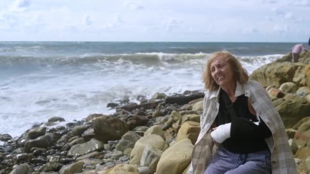 Senior elderly woman relaxing sitting on coast of ocean or sea windy cloudy day suffering from pain. Injury broken arm hand. Old lady wrist in gypsum sling bandage feeling hurt outside in summer — Stock Video