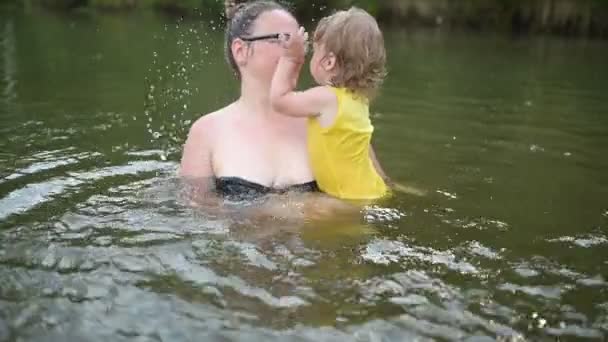 黄色のボディスーツの小さな面白いかわいいブロンドの女の子の子供の幼児笑いは夏の湖で外で泳ぐことを学びます。プラスサイズボディ肯定的な笑顔の女性の母親の赤ちゃんは自然のプールで泳ぐ。水が噴き出す. — ストック動画