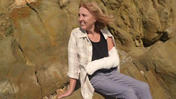 Senior smiling elderly woman relaxing sitting on rocky coastline in windy day suffering from joint pain. Injury broken arm hand. Old lady wrist in gypsum sling bandage feeling hurt outside in summer. — Stock Video