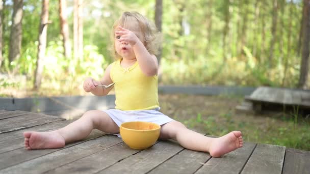 Mały zabawny cute blondynka dziecko maluch z brudnych ubrań i twarzy jedzenia owoców żywności dla dzieci lub puree warzywne z łyżeczką z żółtego talerza na zewnątrz w lecie. Zdrowa koncepcja szczęśliwego dzieciństwa. — Wideo stockowe